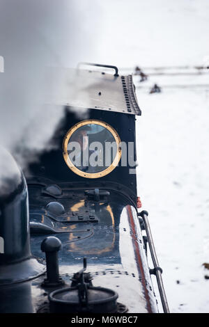 Close front view of steam engine driver inside vintage UK steam locomotive cab, looking through small round train window in the snow. Stock Photo