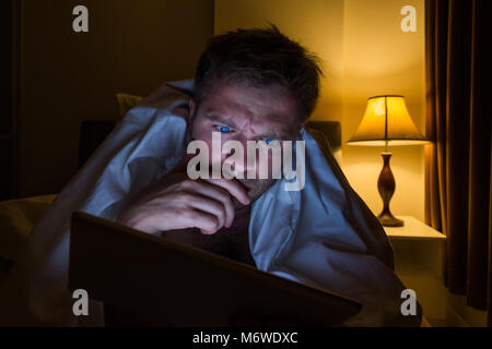 Handsome young man at home reading with tablet PC lying on bed at night. He is tired and want to sleep Stock Photo
