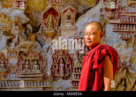 A Buddhist monk is standing in front of rock carvings on the ceiling at Kaw-goon cave, also known as Kawgun Cave Temple or Cave of the Ten Thousand Bu Stock Photo