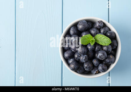 Overhead shot of fresh blueberries piled into a white bowl with a fresh sprig of mint. Set on pale blue, painted wood plank table. Stock Photo
