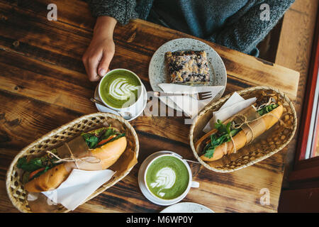 Top view - The girl sitting in cafe and holding mug with green tea with milk next to piece of sweet pie and two sandwiches with vegetables. Stock Photo