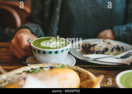 The girl sitting in cafe and holding mug with green tea with milk next to piece of sweet pie and two sandwiches with vegetables. Stock Photo