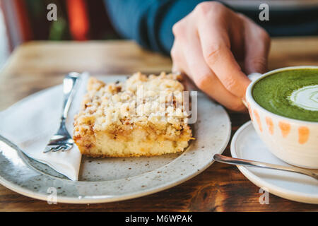 Close-up man's hand holding mug of green tea with beautiful pattern in the form of white foam next to dessert