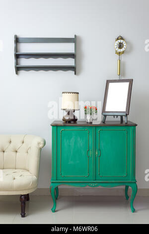 Wooden green vintage sideboard with empty wooden ornate brown desktop photo frame, flower planter, table lamp and cream tub chair on off white wall Stock Photo