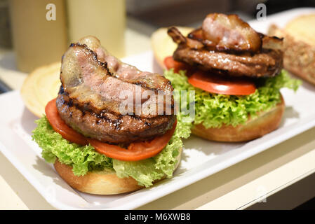 Preparing a hamburger in a restaurant with fresh salad trimmings, beef patty and bacon rashers layered on a white bread roll on a tray Stock Photo