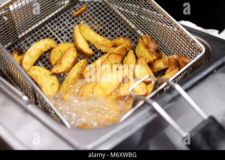 Spicy potato chips or wedges cooking in a deep fryer in a close up view of the hot bubbling oil and wire metal basket Stock Photo