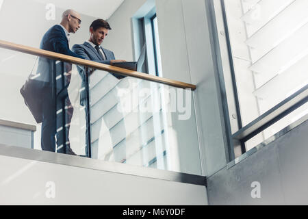 Low angle view of two businessmen looking at laptop while standing by a railing. Business people working together in office. Stock Photo