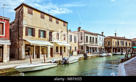 Daylight view to Venetian Lagoon canal with parked boats and tourists walking on sidewalk. Bright blue sky with clouds and historic architecture build Stock Photo