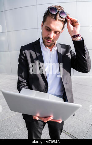Male young handsome surprised businessman or worker in black suit with white shirt in suglasses standing outside and looking into laptop with minimali Stock Photo