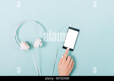 Mobile phone with blank screen in female hand and white headphones on blue table top view Stock Photo