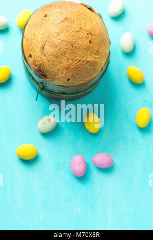 Home Baked Small Easter Cake Panettone with Raisins in Paper Form Multicolored Speckled Chocolate Candy Eggs Scattered on Blue Tabletop. Clean Minimal Stock Photo