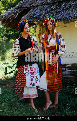 two Beautiful Ukrainian girl  blond and brunette in national embroidered clothes on a background of a wooden hut in summer Stock Photo