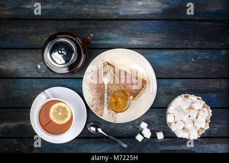 Slice of Cheesecake with jam and tea kettle with lemon and rafinated sugar on a colored wooden background. Top view Stock Photo