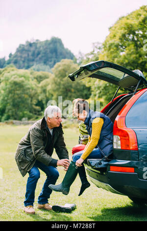 Senior couple have arrived to a national park in their car. The woman is sitting on the edge of the car boot and the man is helping her put her wellie Stock Photo
