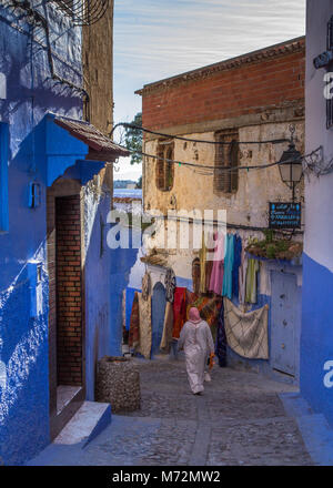 A back view of local woman in traditional dress djeibella walking down colourful narrow street scene in the blue city of Chefchaouen, Morocco Stock Photo