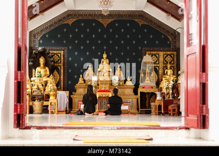 Two women in black clothes sitting in pray inside a temple of the Grand Palace in Bangkok, in front of Buddha statues. Back view from entrance door Stock Photo