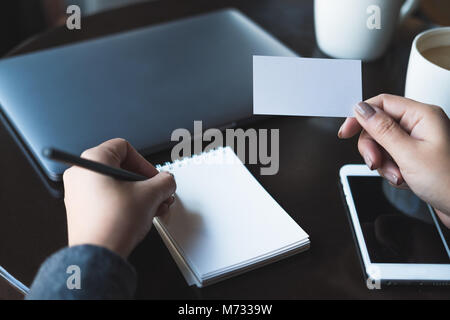 Closeup image of a woman's hands holding empty white business card and writing on blank notebook with laptop and mobile phone on the table Stock Photo