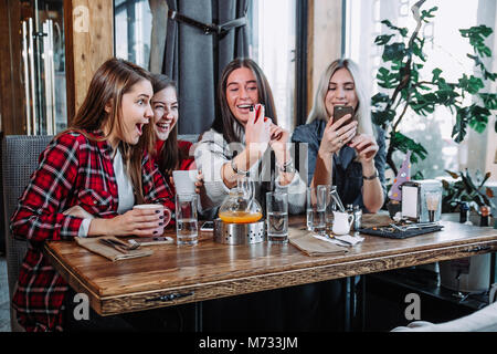 four young women sitting in cafe using smart phone and having funny conversation. Stock Photo
