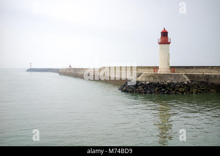 The lighthouse and pier of La Cotiniere port on the Oleron island by a misty morning with still waters under a grey sky. Stock Photo