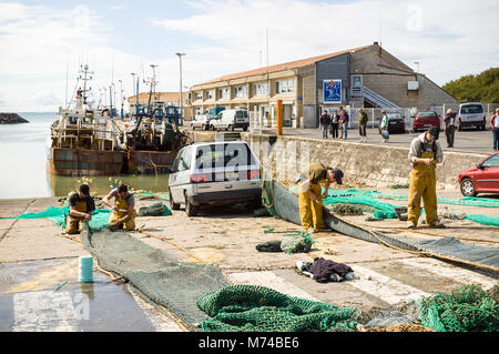 Fishermen mend nets in the port of La Cotiniere on the island of Oleron, France, with fishing boats moored in front of the fish market building. Stock Photo