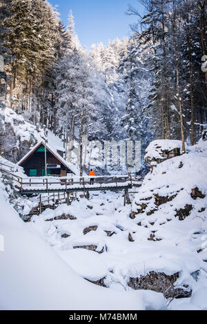 Man with orange jacket standing near wooden hut on bridge in snow covered frozen icy gorge Baerenschuetzklamm and looking to blue sky Stock Photo