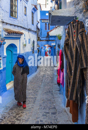 A local man in traditional dress clothing walks along colourful narrow cobbled street in the blue city of Chefchaouen, Morocco. Djebella hanging up. Stock Photo