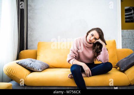 Picture of a tired unkempt woman sitting on he couch at home. Girl watch TV new serial. Sofa relax concept after hard working day. Attentive woman sitting bored on sofa at home Stock Photo