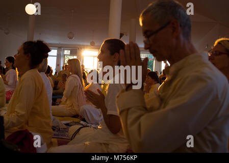 A hall full of people who praying with folded hands and closed eyes while sun is shining through a windows during sunset. People attending summer yoga Stock Photo