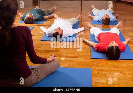 Young people practising relaxation also called yoga nidra. Guided by an instructor laying in shavasana on blue yoga mats on parquet flooring indoors. Stock Photo