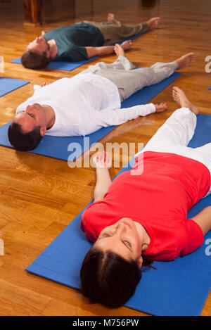 Three young people practising relaxation also called yoga nidra. Laying in shavasana on blue yoga mats on parquet flooring indoors. Stock Photo