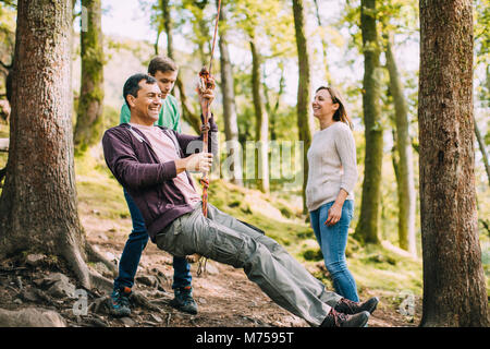 Mature man is being pushed on a rope swing by his son while out on a hike with his family. Stock Photo