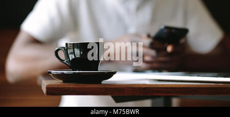 Closeup of a coffee cup placed on a table in a coffee shop. Cropped shot of man sitting at a coffee shop with a glossy black coffee cup and saucer on Stock Photo