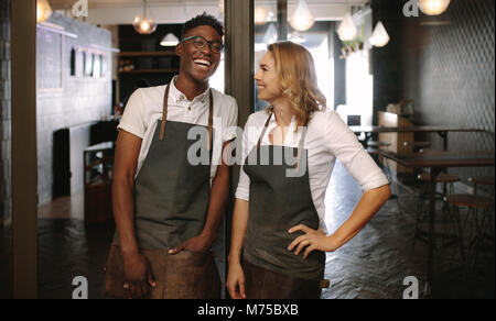 Happy young coffee shop owners standing at their shop. Man and woman baristas standing inside café wearing apron. Stock Photo