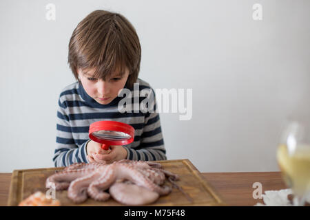 Cute preschool child, boy, analyzing raw octopus with magnifying glass, sitting on a wooden choping board on the table Stock Photo