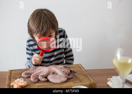 Cute preschool child, boy, analyzing raw octopus with magnifying glass, sitting on a wooden choping board on the table Stock Photo