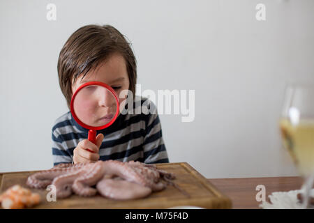 Cute preschool child, boy, analyzing raw octopus with magnifying glass, sitting on a wooden choping board on the table Stock Photo