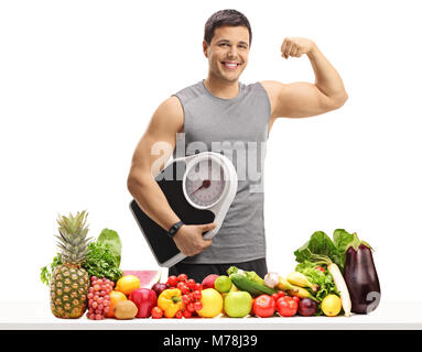 Young man in sportswear holding a weight scale and flexing his biceps behind a table with fruit and vegetables isolated on white background Stock Photo