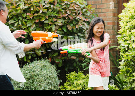 Little girl and her father are having a water fight in the garden with water pistols. The little girl is getting soaked and has her eyes closed as she Stock Photo