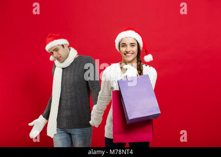 Christmas Concept - Beautiful girlfriend force her boyfriend to go shopping over red christmas background. Stock Photo