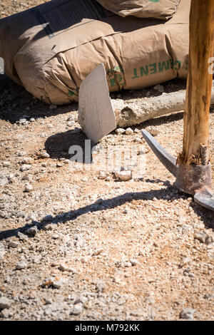 Tools and material used on a Construction Site. Bag of Cement, hoe and pickaxe on the ground outside used by construction workers on hot sunny day. Stock Photo