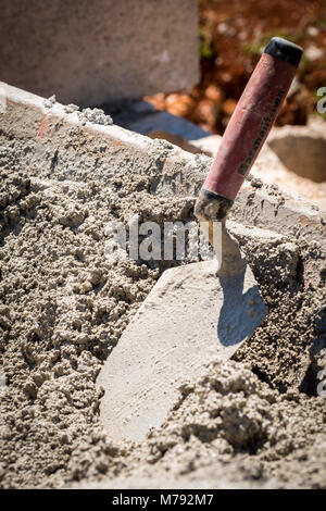 Construction Site with workers using trowel to spread wet cement from wheelbarrow onto cement blocks to build a concrete wall with steel rods Stock Photo