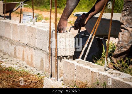 Construction worker on site using trowel to spread wet cement from his wheelbarrow between concrete blocks to hold blocks in place over steel rods Stock Photo