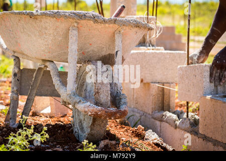 Hands of construction worker laying cement blocks over steel rods building a concrete wall. Wheelbarrow filled with wet cement to hold blocks together Stock Photo