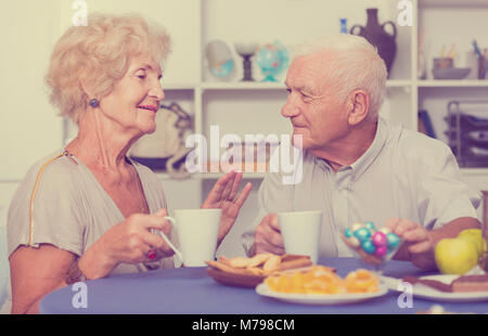 Smiling elderly spouses enjoying time at home, drinking tea with sweets Stock Photo