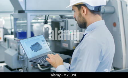 Chief Engineer in the Hard Hat Holds Laptop with 3D Component Model on it's Screen. In the Background Modern Factory Equipment. Stock Photo