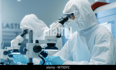 Two Engineers/ Scientists/ Technicians in Sterile Cleanroom Suits  Control Manufacturing Machinery Work and Use Microscopes for Component Adjustment Stock Photo