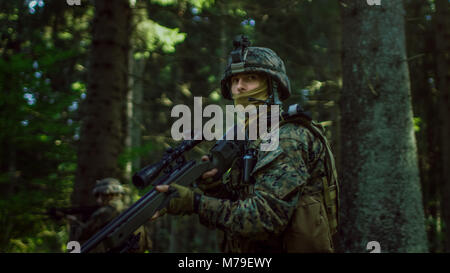 Fully Equipped Sniper Soldier Wearing Camouflage Uniform Attacking Enemy, Rifle in Firing Position. Military Operation in Action in Forest. Stock Photo