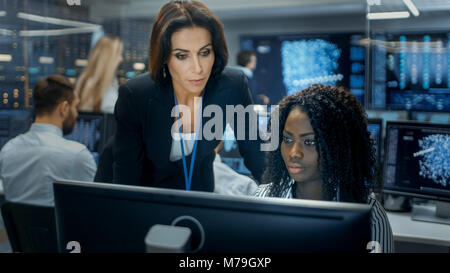 Female Team Leader Consults Young Computer Engineer. They Work in a Crowded Office on a Neural Network/ Artificial Intelligence Project. Stock Photo