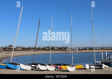 A row of five small colorful catamarans resting on a curving beach, ready for sailing lessons, masts against a blue sea and sky,  copyspace, nobody Stock Photo