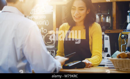 In the Cafe Beautiful Hispanic Woman Makes Takeaway Coffee For a Customer Who Pays by Contactless Mobile Phone to Credit Card System. Stock Photo